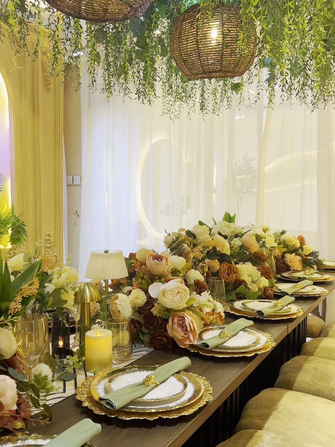 A long dining table set with gold chargers, sage napkins and rose centrepieces beneath hanging greenery and woven pendant lamps